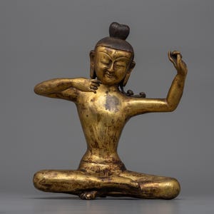 May include: A gold-coloured metal statue of a seated Buddha with a dark brown bun on its head. The Buddha is in a meditative pose with its hands raised in a gesture of blessing.