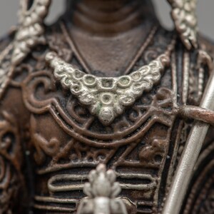 May include: Close-up of a bronze statue of a warrior wearing ornate armor with silver accents. The warrior is holding a spear in their right hand.