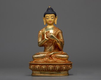 May include: A gold-coloured statue of Buddha sitting in meditation pose with his hands in his lap. The statue is detailed with intricate carvings and is set on a base with a decorative design.