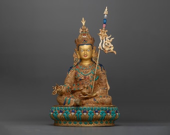 May include: A gold-colored statue of a seated figure with a crown and intricate details. The figure is holding a small object in their right hand and has a serene expression. The statue is set on a base with a lotus design and is adorned with turquoise and other gemstones.