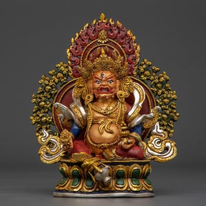 Könnte beinhalten: Eine mehrfarbige Statue einer Gottheit mit grimmigem Ausdruck, drei Augen und einer verzierten Krone. Die Figur sitzt auf einem Lotussockel, hält in jeder Hand einen kleinen Gegenstand und ist von einem detaillierten, flammenartigen Heiligenschein umgeben.