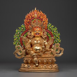 Könnte beinhalten: Goldene Statue einer sitzenden Gottheit mit grimmigem Ausdruck, geschmückt mit Krone und aufwendigem Schmuck. Die Statue steht vor einem Hintergrund aus roten, grünen und goldenen dekorativen Elementen, darunter Flammen und florale Muster. Der Sockel ist mit kunstvollen Schnitzereien versehen.
