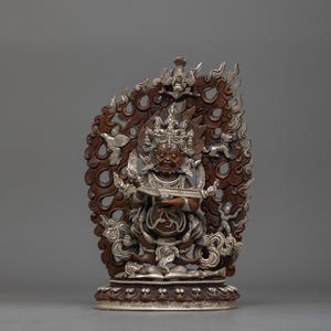 May include: A silver and brown Tibetan deity statue with a fierce expression, holding a sword and a drum. The statue is surrounded by a decorative flame-like halo.