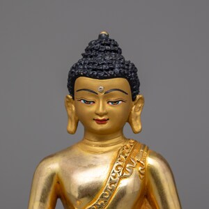 May include: A gold-painted statue of Buddha with a black hairpiece and a small, clear gem on the forehead. The statue is wearing a gold robe with intricate designs.