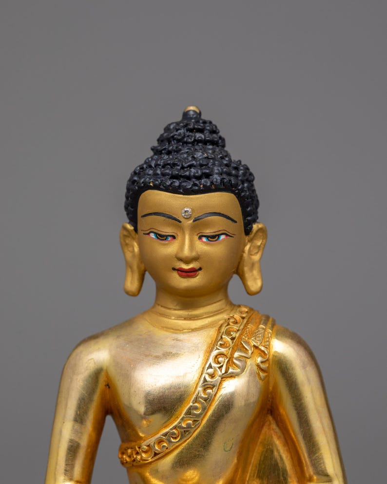 May include: A gold-painted statue of Buddha with a black hairpiece and a small, clear gem on the forehead. The statue is wearing a gold robe with intricate designs.