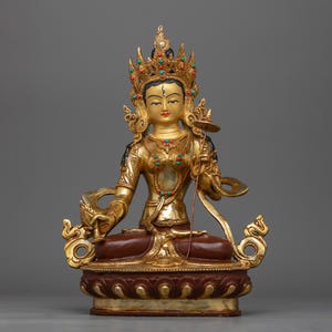 May include: A gold-coloured statue of a deity, sitting in a meditative pose. The statue is adorned with intricate details and colourful gemstones. The deity is wearing a crown and holding a staff.