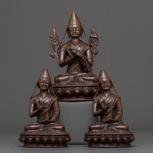 Puede incluir: Tres estatuas de bronce de figuras budistas en posición de meditación sentada. La figura central es más grande y lleva una corona. Las tres figuras tienen tocados elaborados y están adornadas con detalles intrincados.