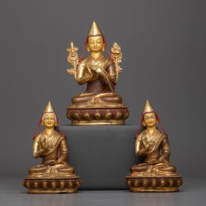 Puede incluir: Tres estatuas doradas y rojas pintadas de figuras budistas que llevan sombreros tradicionales. Las estatuas están sentadas sobre una superficie gris oscura.