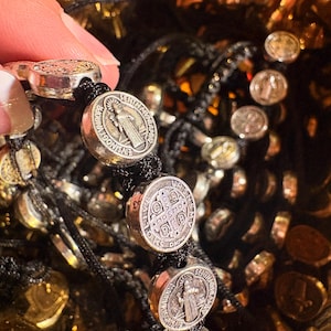 May include: Silver-toned religious bracelet featuring round medallions with detailed engravings and black cord. The medallions display religious figures and symbols. The bracelet is held by a hand, showcasing the intricate design and craftsmanship.