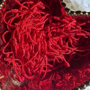 May include: A heart-shaped, red dish filled with tangled red yarn. The dish has a gold-colored trim and a textured surface. The yarn appears to be made of a shiny, silky material.