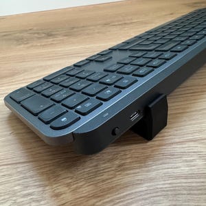 Op de afbeelding: Een donkergrijze draadloze toetsenbord met een volledige indeling, inclusief numeriek toetsenbord en functietoetsen. Het toetsenbord heeft een plat ontwerp met een zwarte USB-ontvanger aan de zijkant. De toetsen zijn plat en gelijkmatig verdeeld.