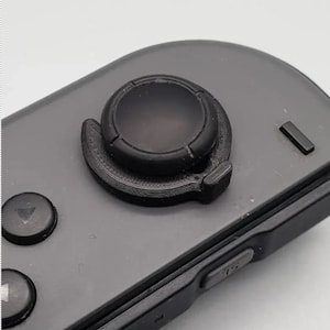 Op de afbeelding: Close-up van een zwarte joystick duimgreep bevestigd aan een grijze gamecontroller. De duimgreep is rond met een textuur en een kleine, gebogen verlenging. De controller heeft verschillende knoppen en een gladde, matte afwerking.