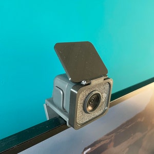 May include: A grey Logitech webcam with a black privacy cover is mounted on a computer monitor. The webcam has a square design with a lens and the Logitech logo. The monitor displays the time and date.