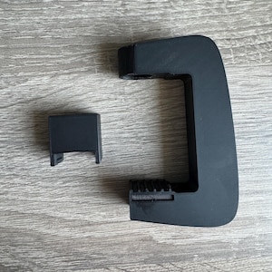 Op de afbeelding: Zwart 3D-geprint object, mogelijk een handvat of steun, met een rechthoekige basis en een gebogen arm. Een apart, kleiner rechthoekig stuk is ook aanwezig. De objecten liggen op een houten oppervlak.