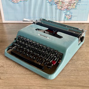 May include: A light blue Olivetti Lettera 22 portable typewriter with a QWERTY keyboard layout, black circular keys, a long black space bar, and a single red key. The metal casing features the brand name and model text clearly printed on the front.