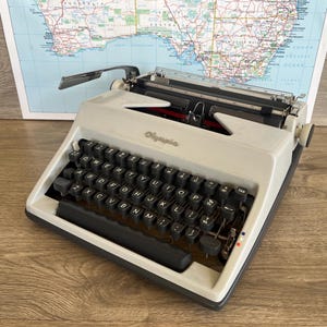May include: A vintage Olympia typewriter in light grey with black keys. The typewriter is on a wooden surface in front of a map of Australia. The Olympia logo is visible on the front.