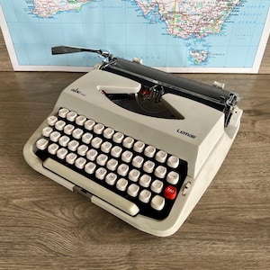 May include: A vintage off-white Lemair typewriter with black keys and a red "TAB" key. The typewriter sits on a wooden surface with a map of Australia in the background. The word "abc" is visible on the top left.