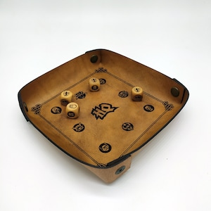 May include: A square, tan leather dice tray with rounded corners and a black border design. Six wooden dice are inside the tray. The tray has a black logo and decorative corner designs.