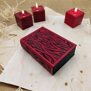 May include: A red and black journal with a zebra stripe pattern sits on a piece of parchment paper. Three square, burgundy candles with lit flames are in the background. The journal has a black spine and edges.