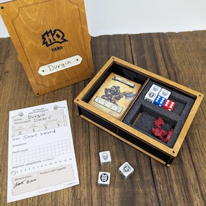 May include: Wooden game box with the word "HERO" and the name "Durgin" on the lid. Inside, a card, dice, and a red figurine are visible. A character sheet and additional dice are also present, all on a wooden surface.