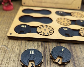May include: A wooden box with a black and gold design, containing circular game pieces with numbers. The box has the word "Trackers" printed on the top. Two game pieces are shown outside the box, one with the number "1" and the other with the number "2".