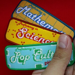Op de afbeelding: Drie kleurrijke stoffen patches met een retro ontwerp. Elke patch heeft een witte rand en de tekst "Proficiency in" gevolgd door een onderwerp: "Mathematics", "Science" en "Pop Culture".