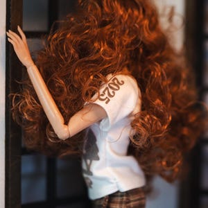 May include: A doll with long, curly, auburn hair wearing a white t-shirt with the number "2023" printed on it and brown plaid pants.