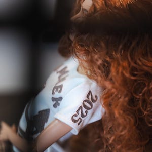 May include: A white t-shirt with the text "2023" and "Miek" printed on it. The shirt is being worn by a doll with long, curly red hair.