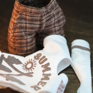 May include: A white onesie with brown text that says "Summer Camp" and a graphic of a tent and sun. There are also two white socks with brown stripes.