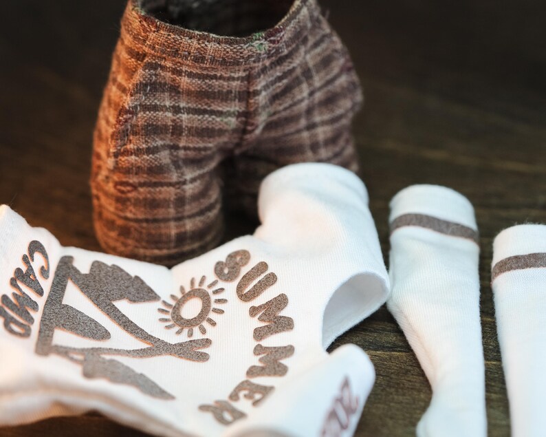 May include: A white onesie with brown text that says "Summer Camp" and a graphic of a tent and sun. There are also two white socks with brown stripes.