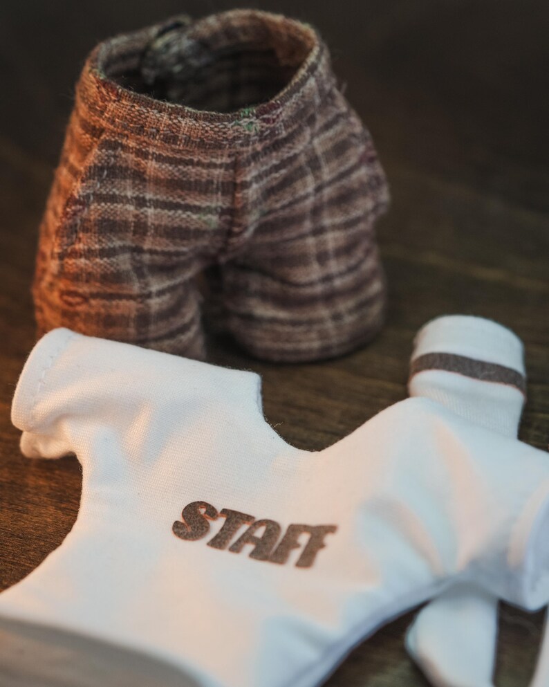May include: A white t-shirt with the word "STAFF" printed on the front, brown and white plaid shorts, and white socks with a brown stripe.
