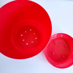 May include: A bright red plastic flower pot with drainage holes in the bottom. The pot is separated into two pieces, a larger bottom piece and a smaller top piece.