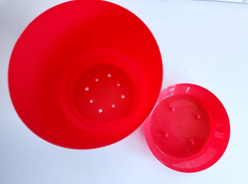 May include: A bright red plastic flower pot with drainage holes in the bottom. The pot is separated into two pieces, a larger bottom piece and a smaller top piece.