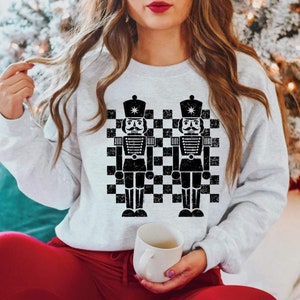 May include: A light gray sweatshirt featuring a black and white checkered pattern with two black nutcracker soldier graphics. The nutcrackers have white mustaches and are wearing black hats. The sweatshirt is a crew neck style.