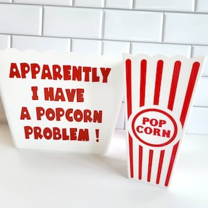 May include: Two white popcorn containers with red accents. One container has the text "APPARENTLY I HAVE A POPCORN PROBLEM!" printed on it. The other container has the text "POP CORN" printed on it.
