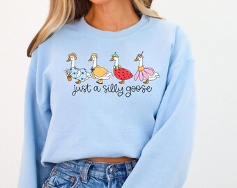 Peut inclure: Un sweat-shirt bleu clair avec un motif de quatre oies portant des tenues différentes. Les oies sont alignées en rangée et le texte "Just a silly goose" est imprimé en dessous.