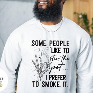 May include: White sweatshirt with a black graphic print of a skeleton hand holding a joint. The text reads "Some people like to stir the pot... I prefer to smoke it."