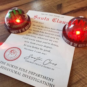 May include: Two red-glowing Santa Cam surveillance devices with the text "PROPERTY OF THE NORTH POLE" and "SANTA CAM". A letter from Santa Claus is also visible, with the text "THE NORTH POLE DEPARTMENT OF BEHAVIORAL INVESTIGATIONS".
