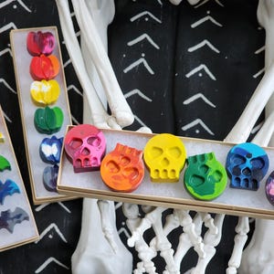 May include: A set of six colorful crayon skulls in a cardboard box. The crayons are arranged in a row and are all different colors, including red, orange, yellow, green, blue, and purple.