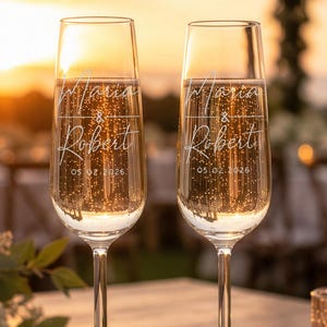 May include: Two clear glass champagne flutes featuring white etched text that reads Maria & Robert and the date 05.02.2026. The tall, slender glasses have a narrow bowl and a long, thin stem, standing upright on a wooden surface.