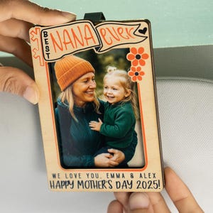 May include: A wooden photo frame with a picture of a woman and a child. The frame has the text "Best Nana Ever" and "We love you, Emma & Alex Happy Mother's Day 2025!" printed on it.