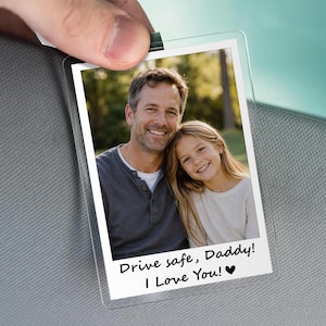 May include: Clear plastic photo holder with a picture of a man and a girl. The photo has the text "Drive safe, Daddy! I Love You!" in black. The holder is being held by a hand.