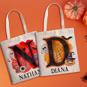 May include: Two white canvas tote bags with Halloween-themed designs. The bag on the left has a black letter N with a spiderweb, an eye, and a ghost. The bag on the right has a black letter D with a spiderweb and a pumpkin. The bags are personalised with the names "Nathan" and "Diana".