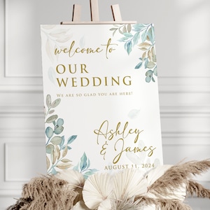 May include: A wedding welcome sign with a white background and gold lettering. The sign features a watercolor design of blue and brown leaves. The text on the sign reads "Welcome to our wedding. We are so glad you are here! Ashley & James August 11, 2024."