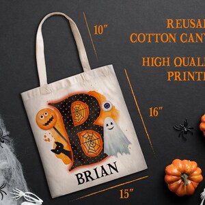 May include: A reusable cotton canvas tote bag with a black and orange Halloween-themed design featuring the letter "B" with a spiderweb, a ghost, a lollipop with a jack-o-lantern face, and a black cat. The bag is personalised with the name "BRIAN" in black lettering. The bag measures 40.6 cm by 38.1 cm.