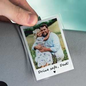 May include: Clear plastic photo holder with a picture of a man and a young girl hugging. The photo has the text "Drive safe, Dad!" and a heart symbol. The holder is being held up.