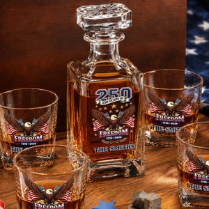 May include: A glass decanter and four matching whiskey glasses filled with amber liquid, featuring a bald eagle graphic, American flags, and the text 250 YEARS OF FREEDOM 1776-2026 THE SMITHS. Includes a wooden box and stone whiskey cubes.