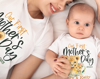 May include: A white t-shirt with the text "Our First Mother's Day 2023" in green and orange lettering. A lion cub and a lioness are pictured in the design. A baby is wearing a matching white onesie with the same design and the text "Our First Mother's Day 2023" in green and orange lettering. The text "Mary & Kelly" is printed below the design.