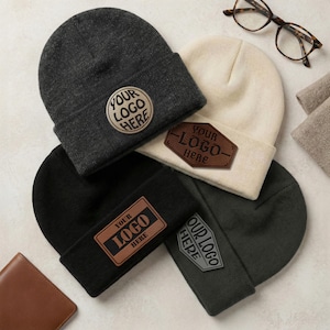May include: Four knit beanies in charcoal, cream, black, and dark green, each featuring a unique leather-like patch with the text "YOUR LOGO HERE" in circular, hexagonal, rectangular, and shield shapes, accompanied by tortoise-shell eyeglasses.