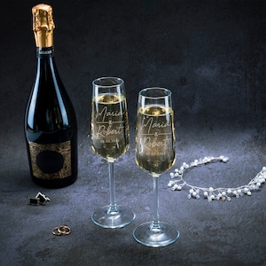 May include: Two champagne flutes with the names Maria and Robert engraved on them, along with the date 09.02.2023. A bottle of champagne and a silver headband with white beads are also in the image.